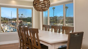 A dining area with a large table and six chairs, large windows with a view of a marina and buildings, and a unique woven pendant light. - Oceanfront dining, shopping, Pool, spa, and sauna on site!- The Harbor Penthouse (Oceanside)
