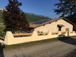 Un edificio rústico con techo inclinado y paneles solares, rodeado por un camino de grava y una valla de madera.