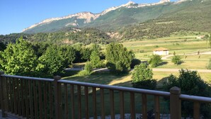Una terraza de madera con vistas a un paisaje verde, que incluye árboles y un edificio a lo lejos.