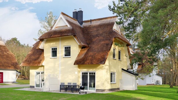 A two-story house with a steeply pitched roof, a front porch with a table and chairs, and a garage attached to the right side.