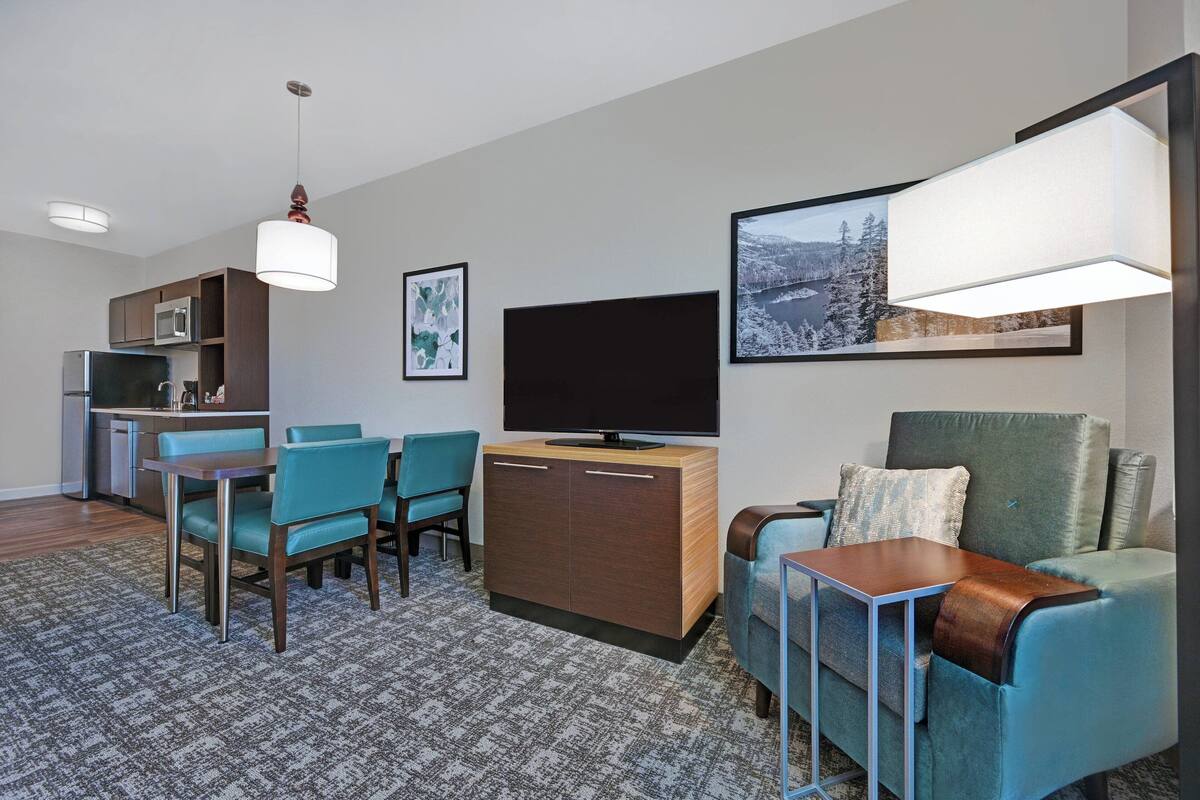 A modern living room with a sofa, a TV, a dining area with chairs, a kitchen area, and a patterned carpet.