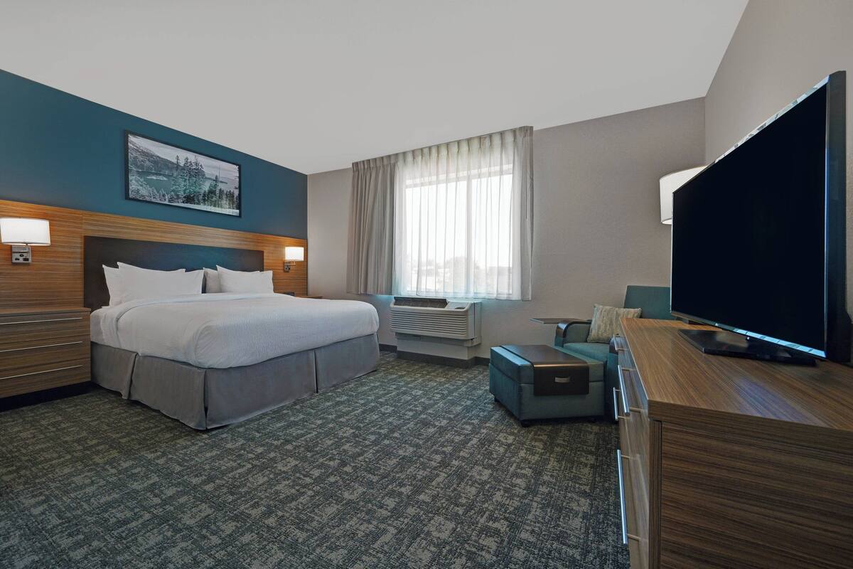 A hotel room with a bed, a desk, a TV, a window with curtains, and an air conditioning unit.