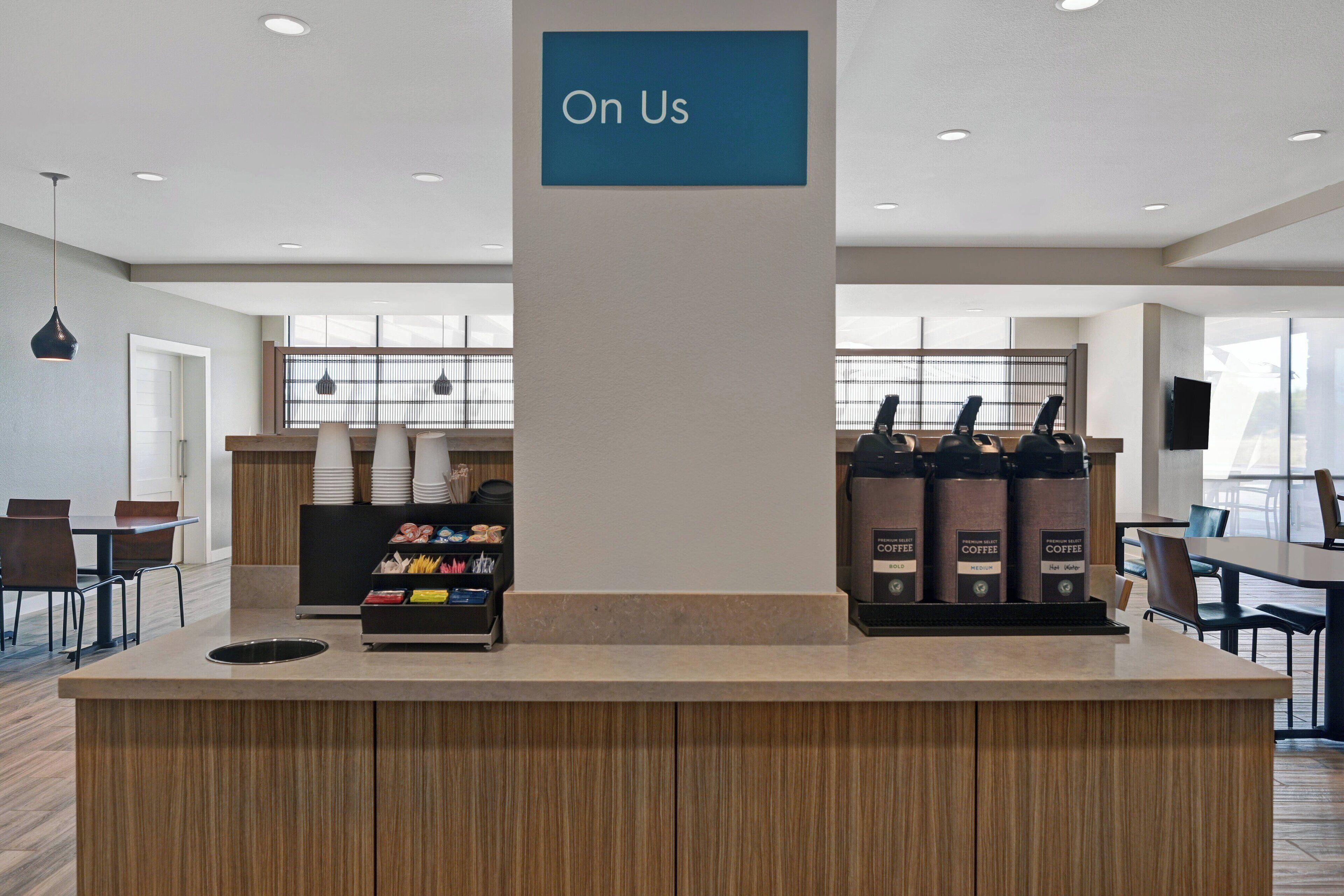 Uma estação de café moderna com um letreiro indicando “Na Conta da Casa” acima do balcão, uma máquina de café e diversos recipientes com café.