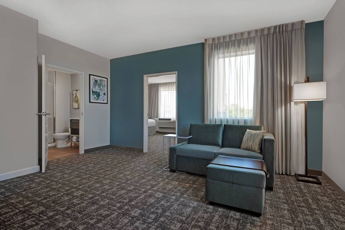 A modern living room with a grey sofa, a small table, and a lamp. There is a door leading to a bathroom and a window with curtains.