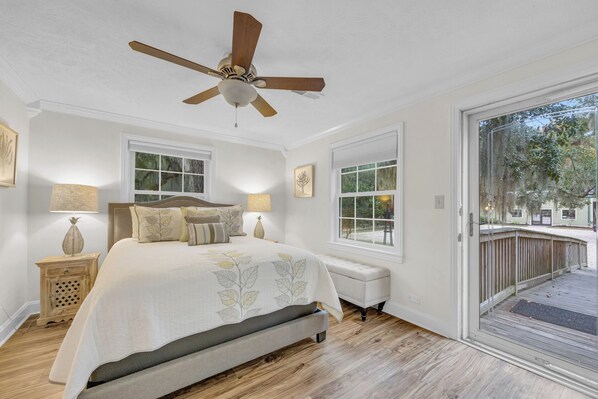 A bedroom with a bed, a ceiling fan, a nightstand, a bench, and a large window.
