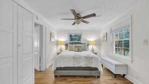 A bedroom with a bed, a ceiling fan, bedside lamps, a bench, and a window with a view of trees.