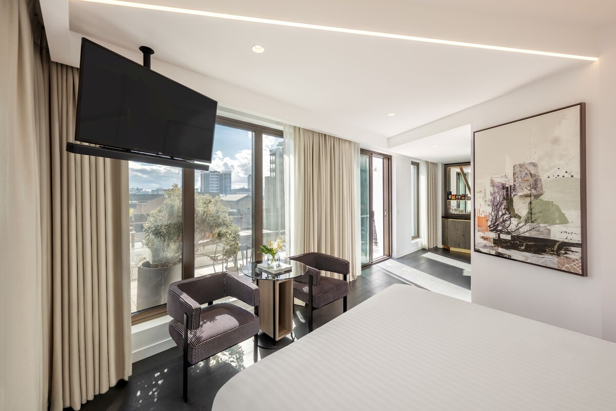 Una habitación de hotel moderna con una cama grande, un televisor de pantalla plana, una zona de comedor con mesa y sillas, y un ventanal con vistas a la ciudad.