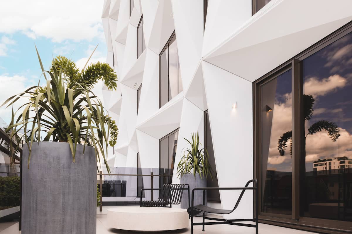 Een modern gebouw met een geometrisch gevelontwerp, grote ramen en een terras met stoelen en plantenpotten.