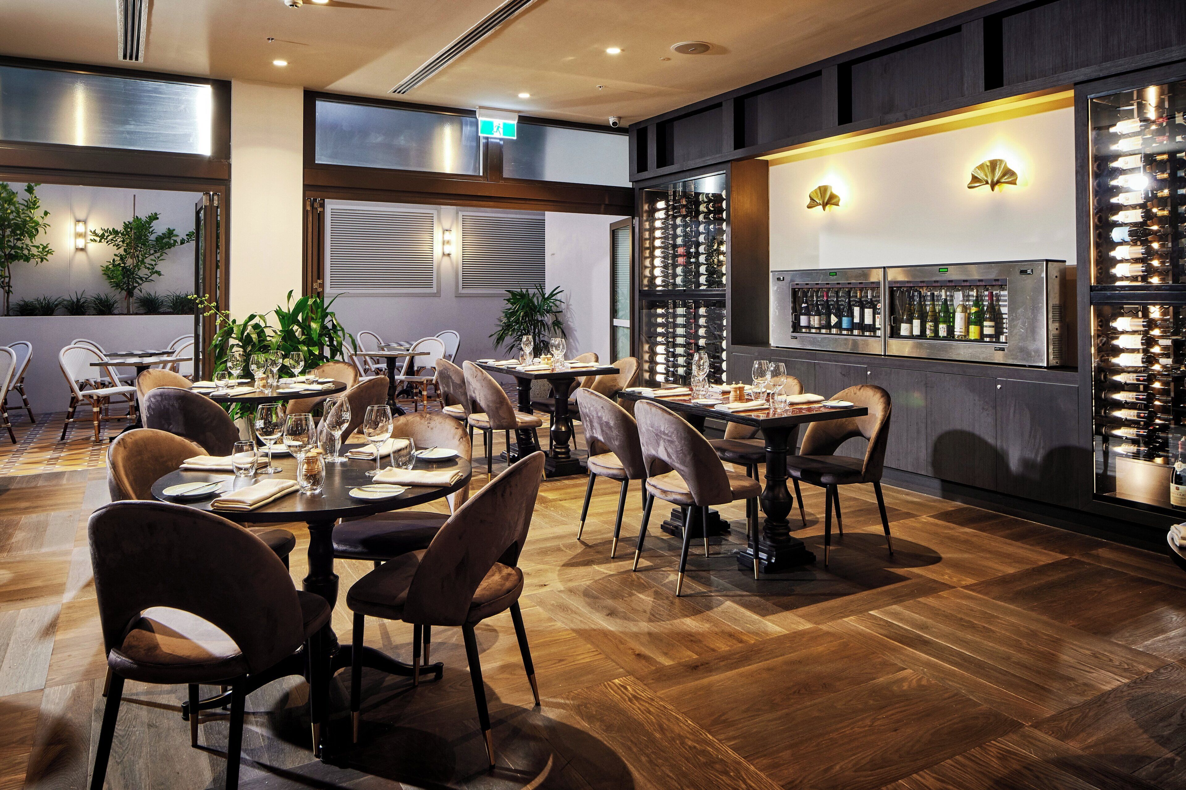 A restaurant with wooden flooring, multiple tables and chairs, a wine refrigerator, and potted plants.