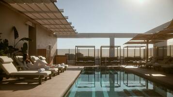 A rooftop pool area with sun loungers and umbrellas, a clear sky, and a view of the cityscape.