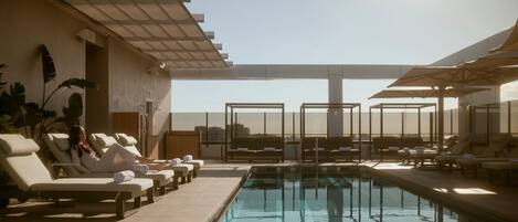 A rooftop pool area with sun loungers and umbrellas, a clear sky, and a view of the cityscape.