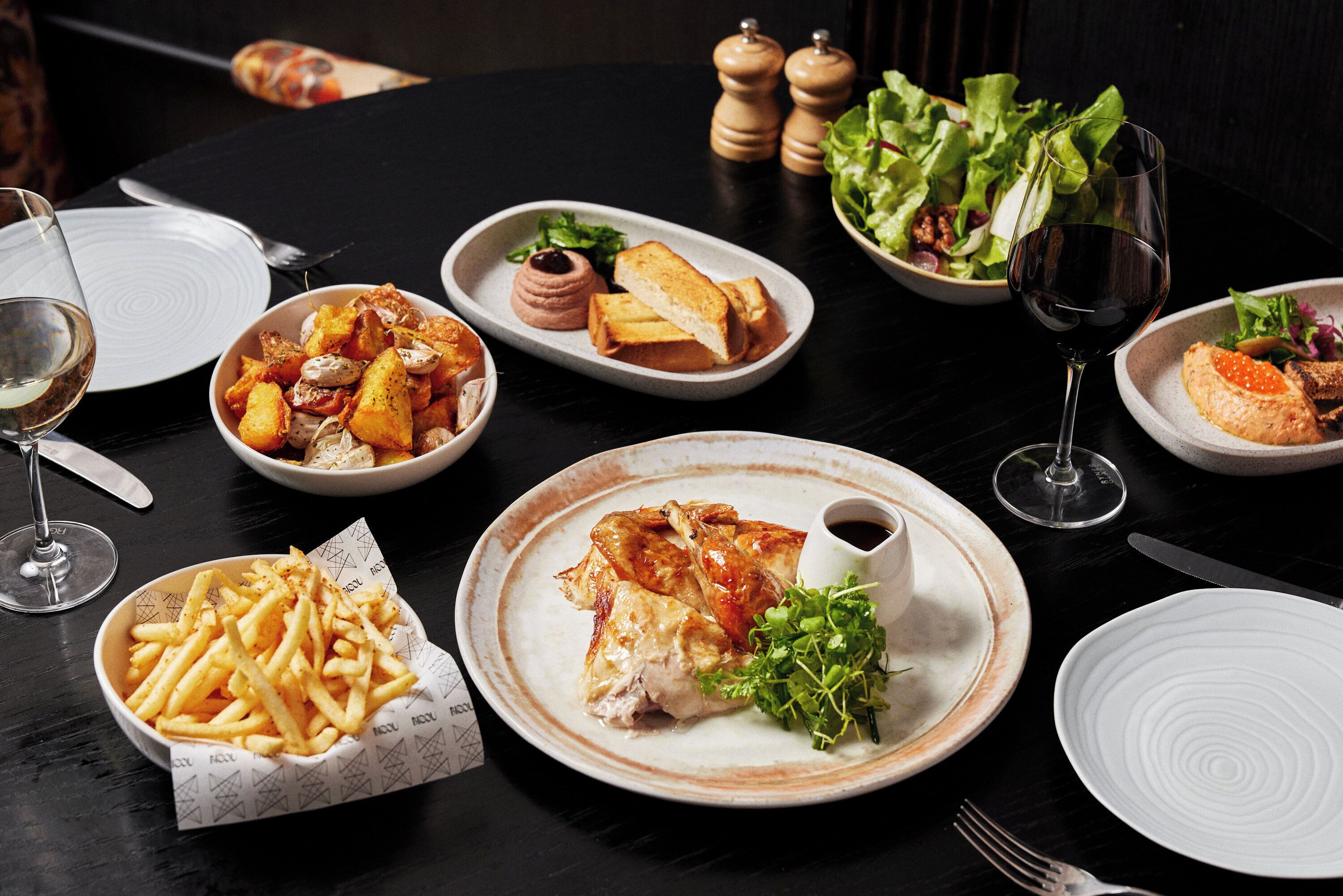 A dining table with various dishes, including a bowl of fries, a plate of chicken, and a glass of wine.