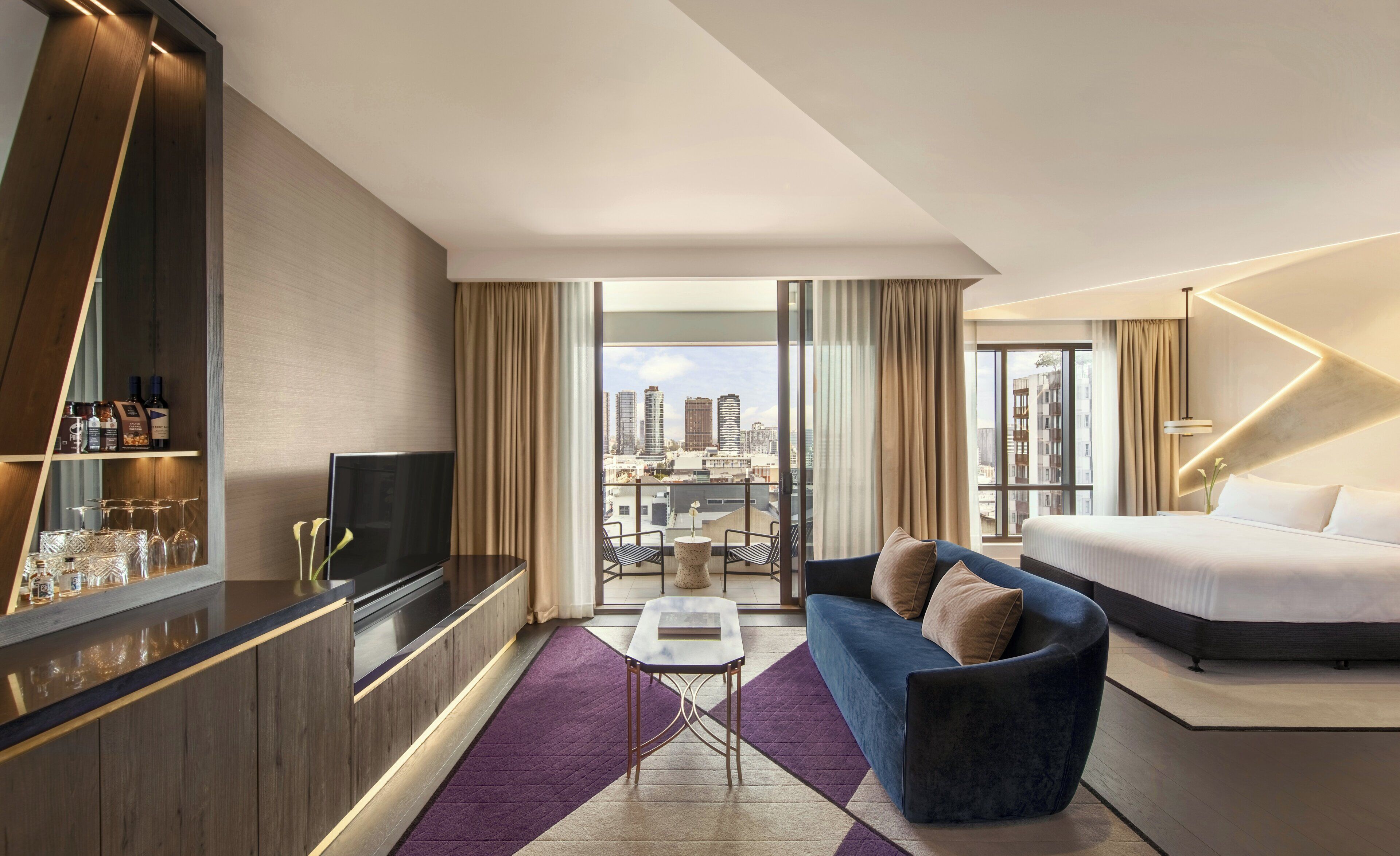 A modern hotel room with a city view, a sofa, a bed, a TV, a minibar, and a balcony with a table and chairs.