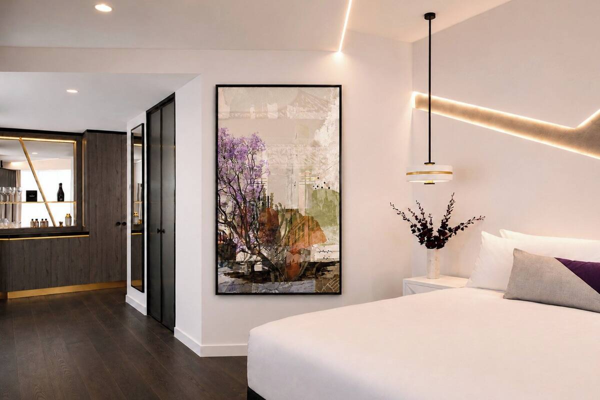 Een moderne hotelkamer met een groot bed, een nachtkastje met een bloevas, een aan de muur hangend kunstwerk en een kitchenette met houten kastjes en een bar.