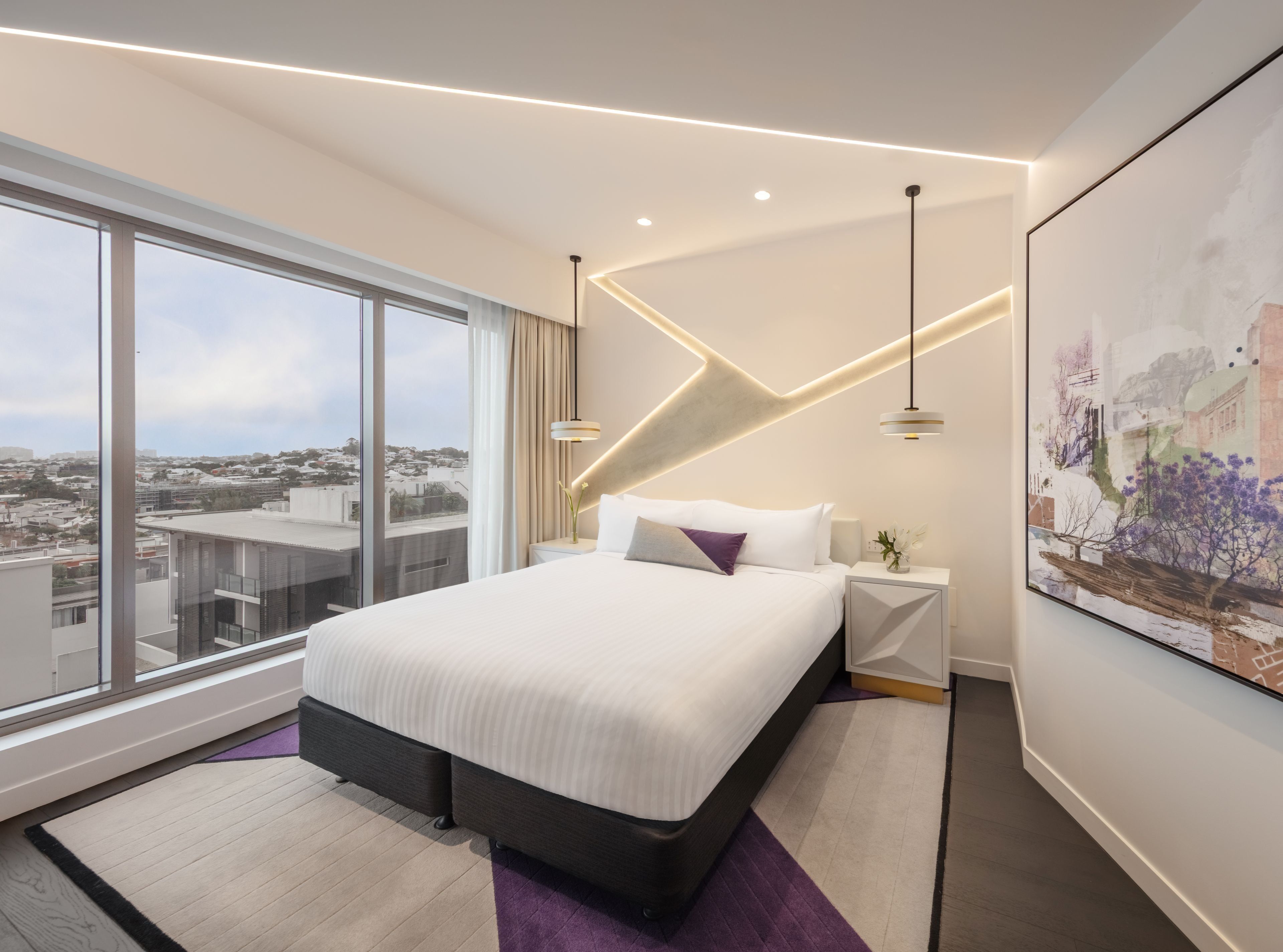 A modern hotel room with a large bed, a city view, a painting on the wall, and a geometric light fixture.