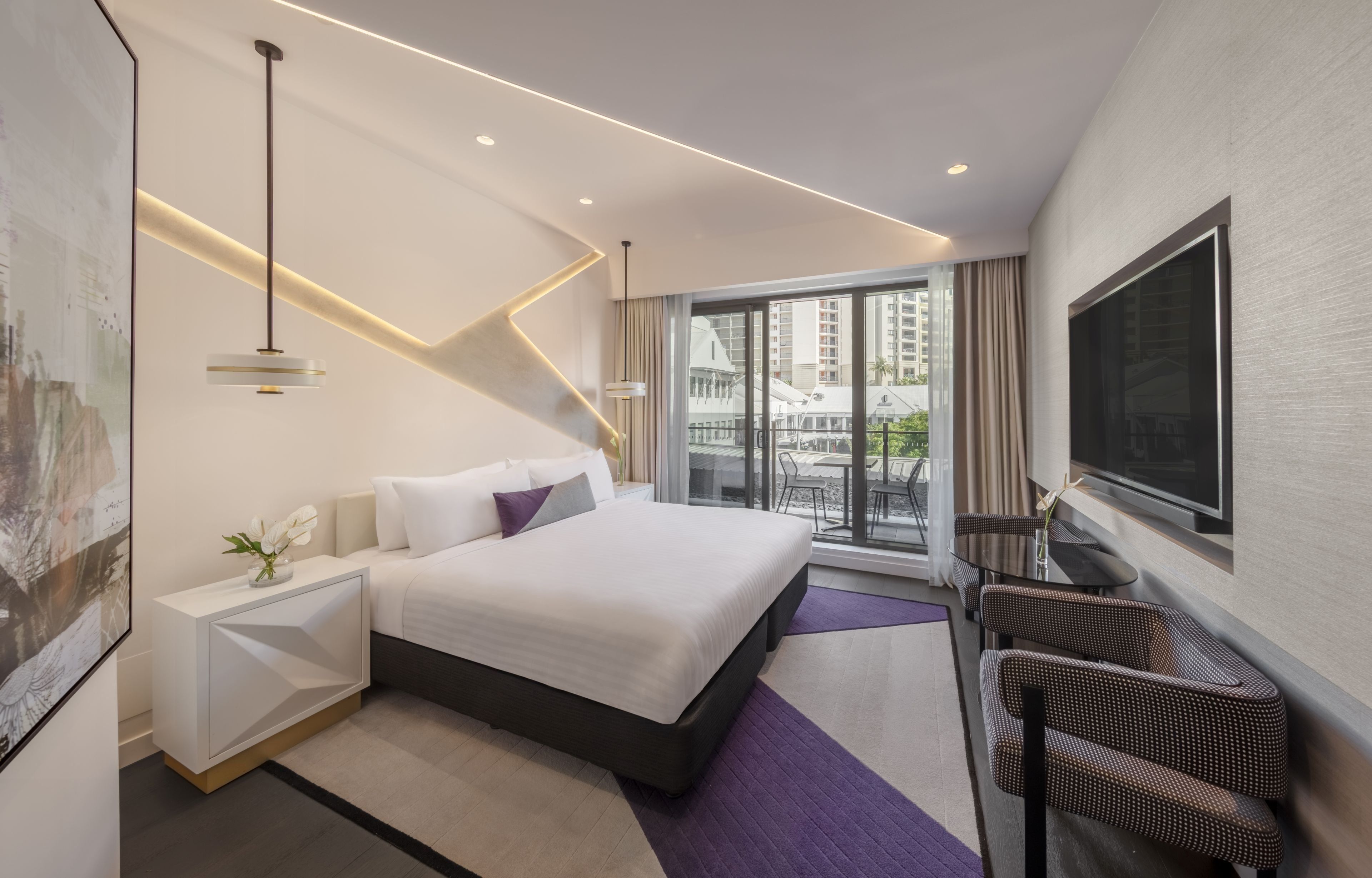 Habitación de hotel moderna con una cama grande, televisión, zona de estar con sillones y vistas a la ciudad a través de amplios ventanales.