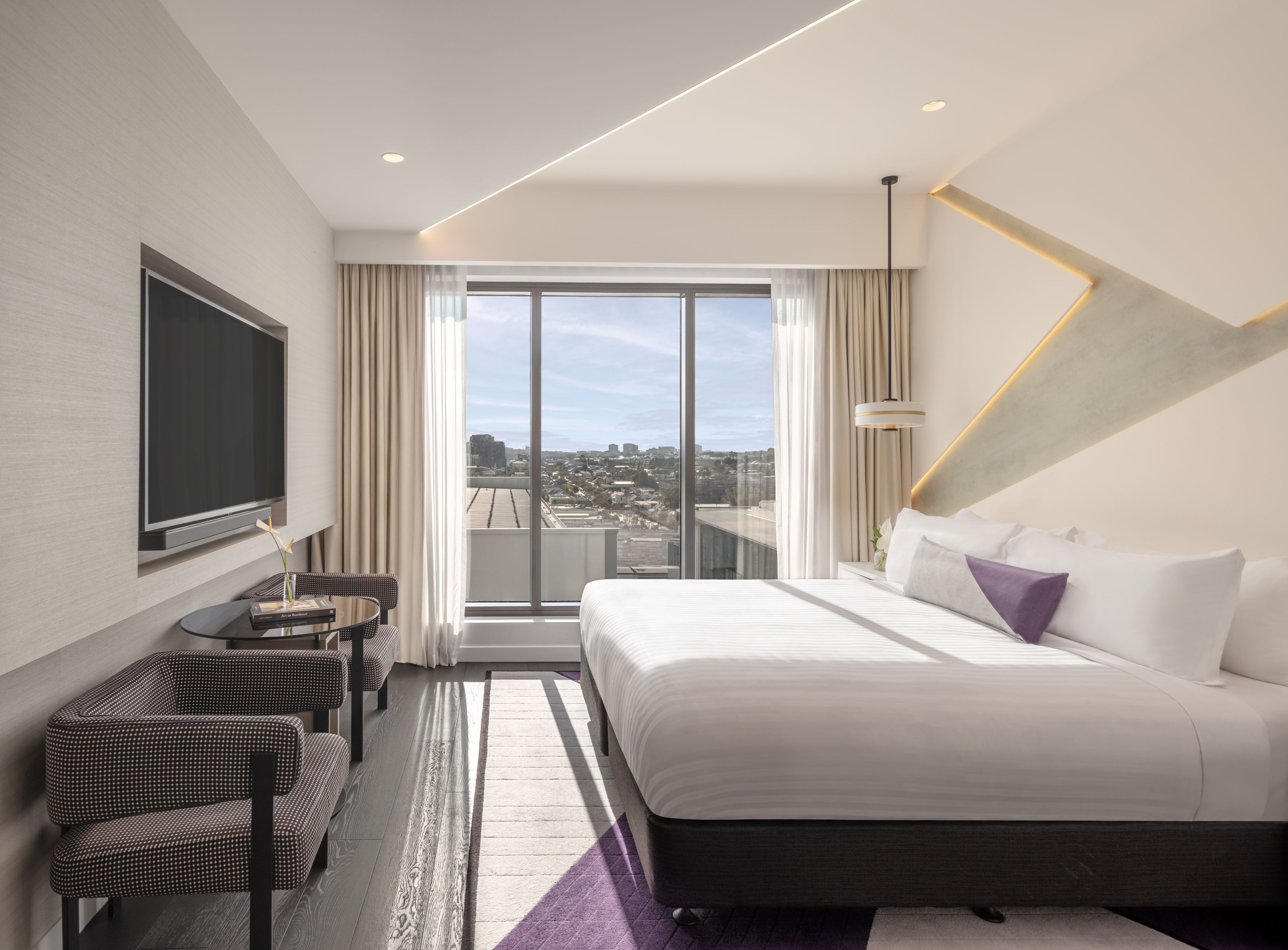 Quarto de hotel moderno com uma cama grande, área de estar com uma mesa e vista para a cidade pela janela.