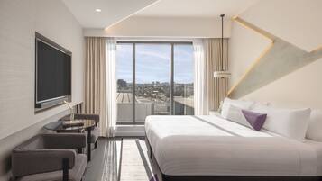 A modern hotel room with a large bed, a seating area with a table, and a city view through the window.
