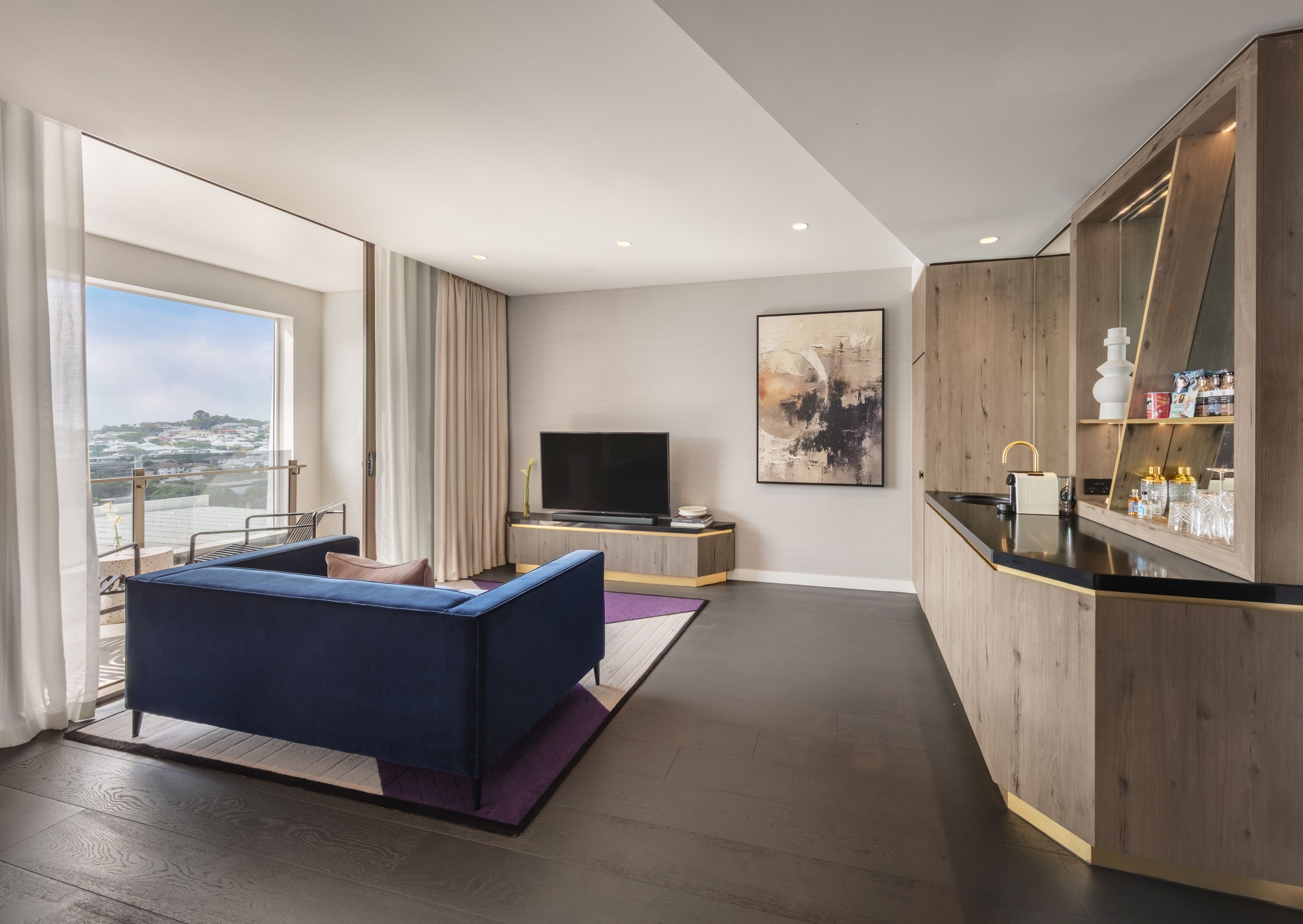 Una habitación de hotel moderna con un ventanal grande, un sofá, un televisor de pantalla plana, una zona de cocina y vistas a la ciudad.