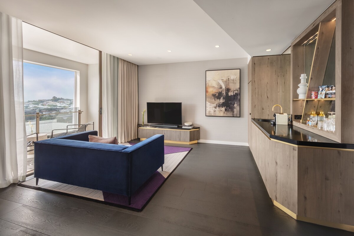 Una habitación de hotel moderna con un ventanal grande, un sofá, un televisor de pantalla plana, una zona de cocina y vistas a la ciudad.