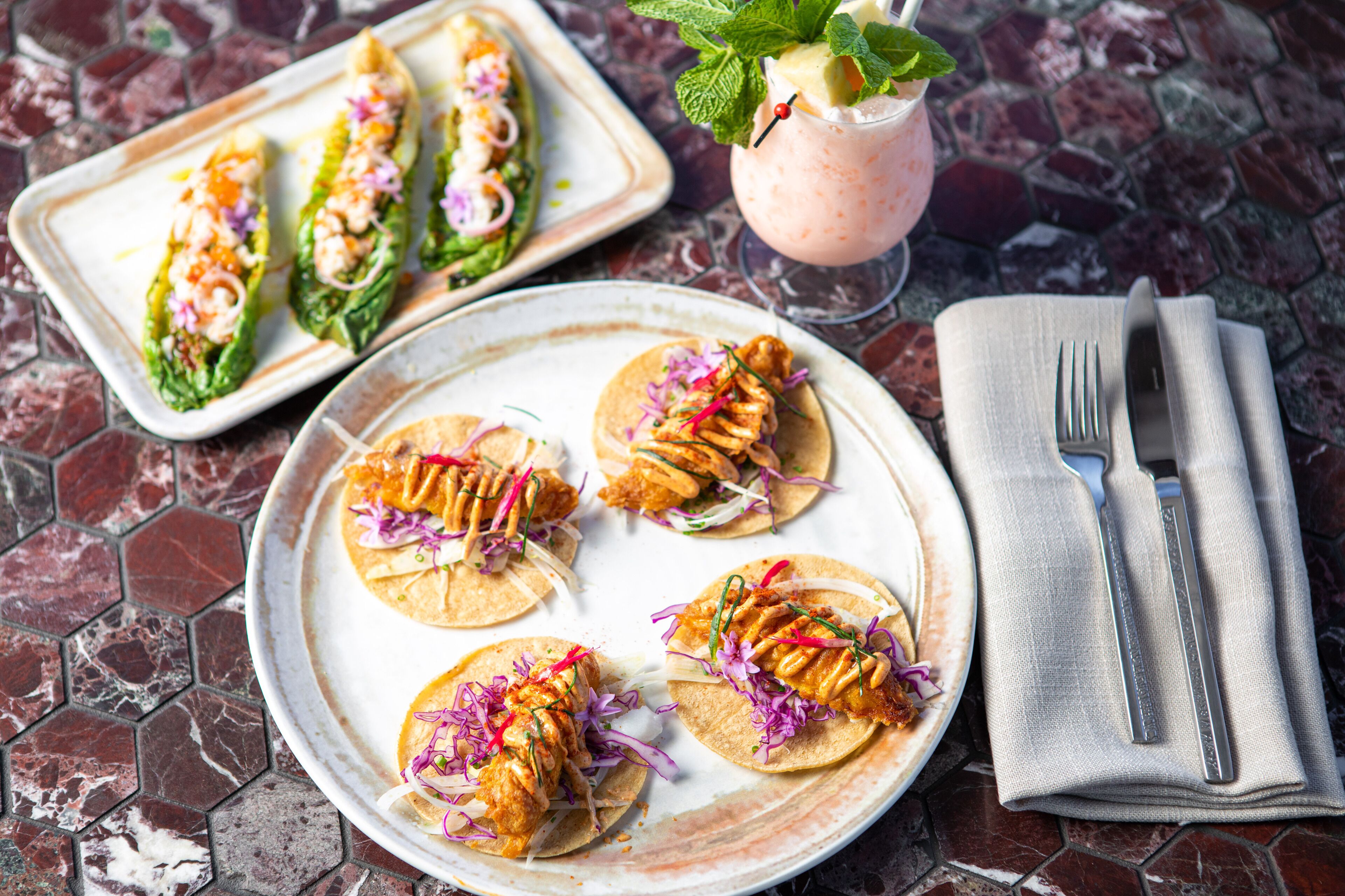 A plate of tacos with a drink and utensils on a tiled table.