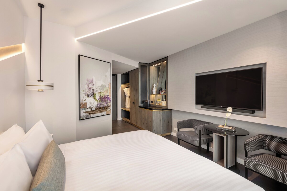 Habitación de hotel moderna con una cama grande, un área de descanso con dos sillas, un televisor de pantalla plana en la pared y una mesita con un florero.