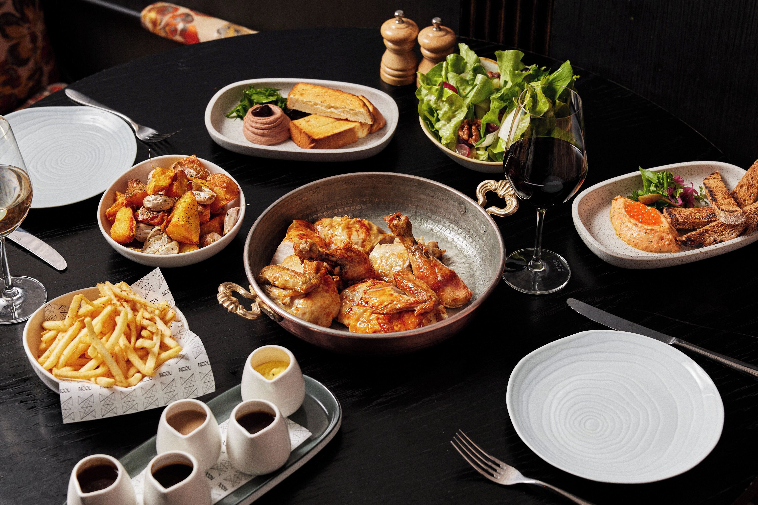 A table set with various dishes including roasted chicken, fries, and salads, accompanied by wine glasses and cutlery.