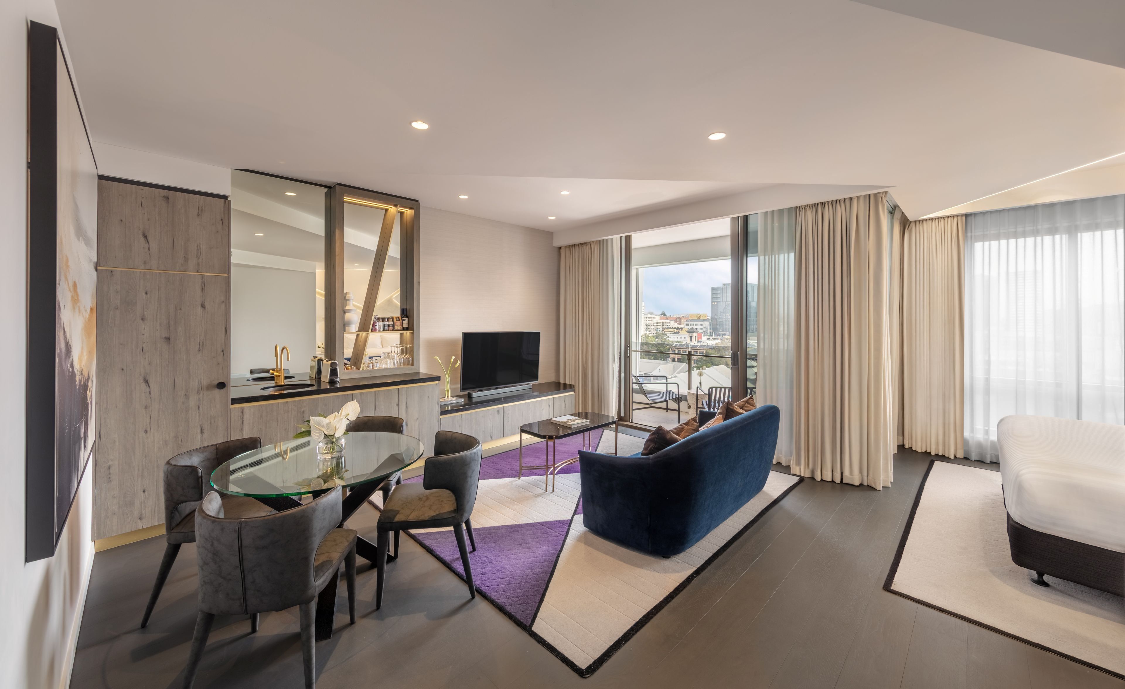A modern hotel room with a dining area, a sofa, a TV, and a balcony with a city view.