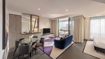 A modern hotel room with a dining area, a sofa, a TV, and a balcony with a city view.