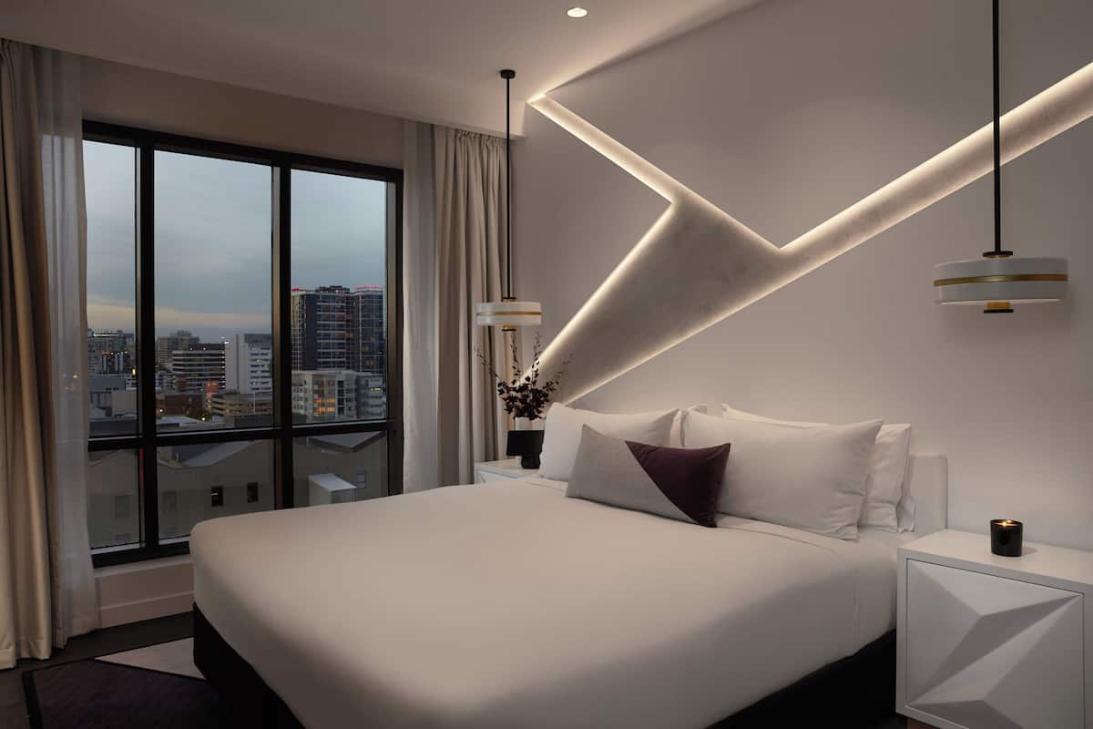 Een moderne hotelkamer met een groot bed, een nachtkastje, een lamp en uitzicht op de stad door grote ramen.