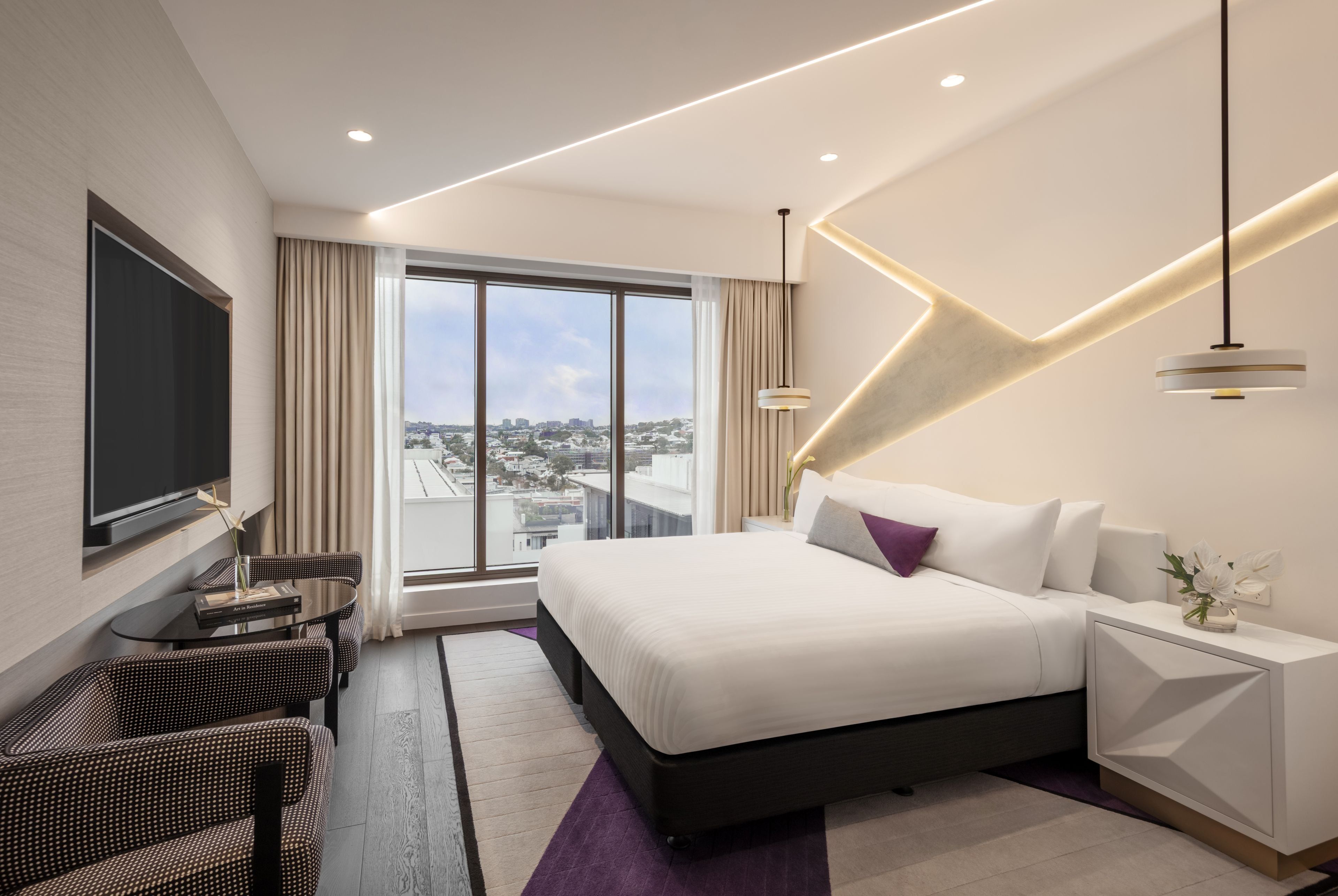 A modern hotel room with a large bed, a flat-screen TV, a desk with a chair, and a view of the city.