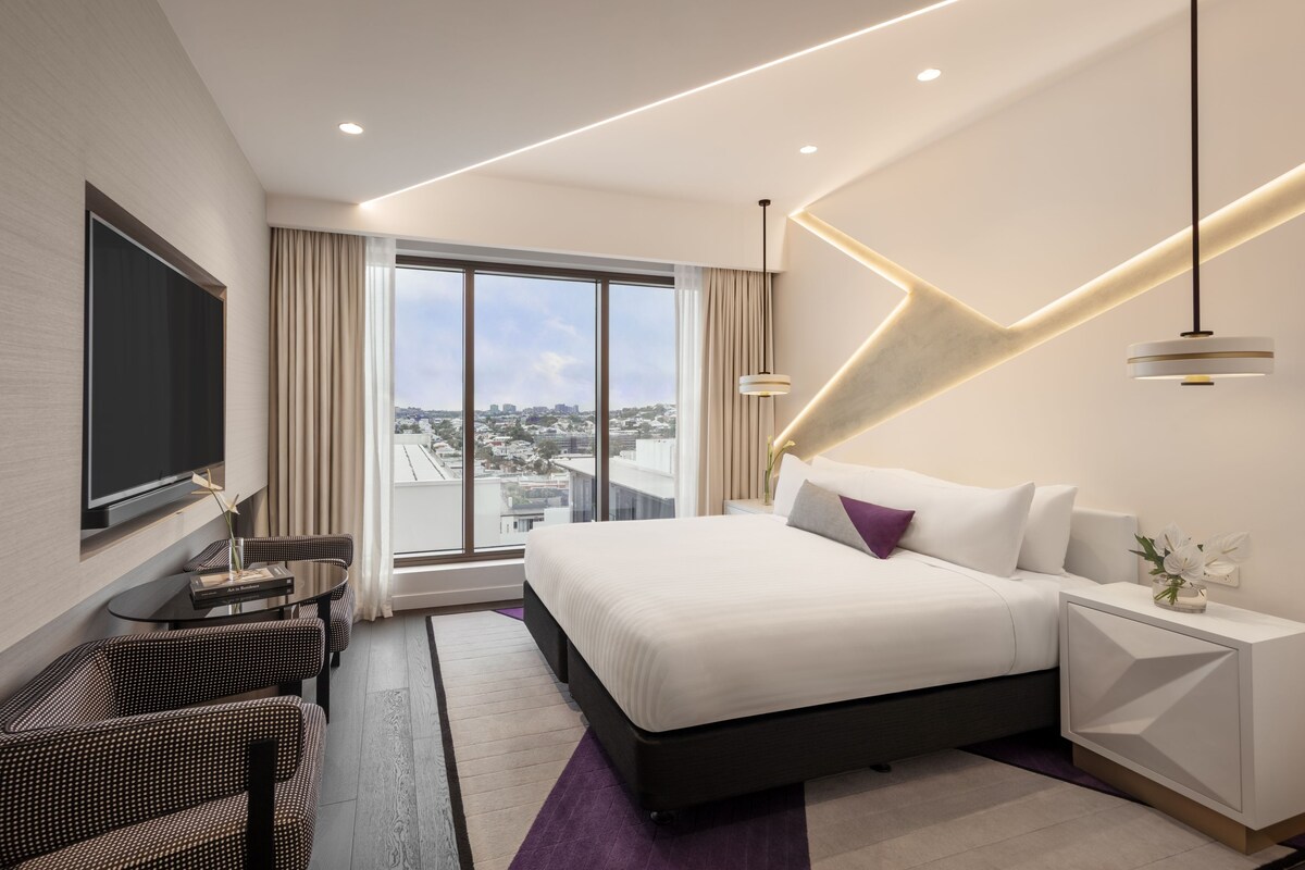 Una habitación de hotel moderna con una cama grande, un televisor de pantalla plana, un escritorio con una silla y vistas a la ciudad.