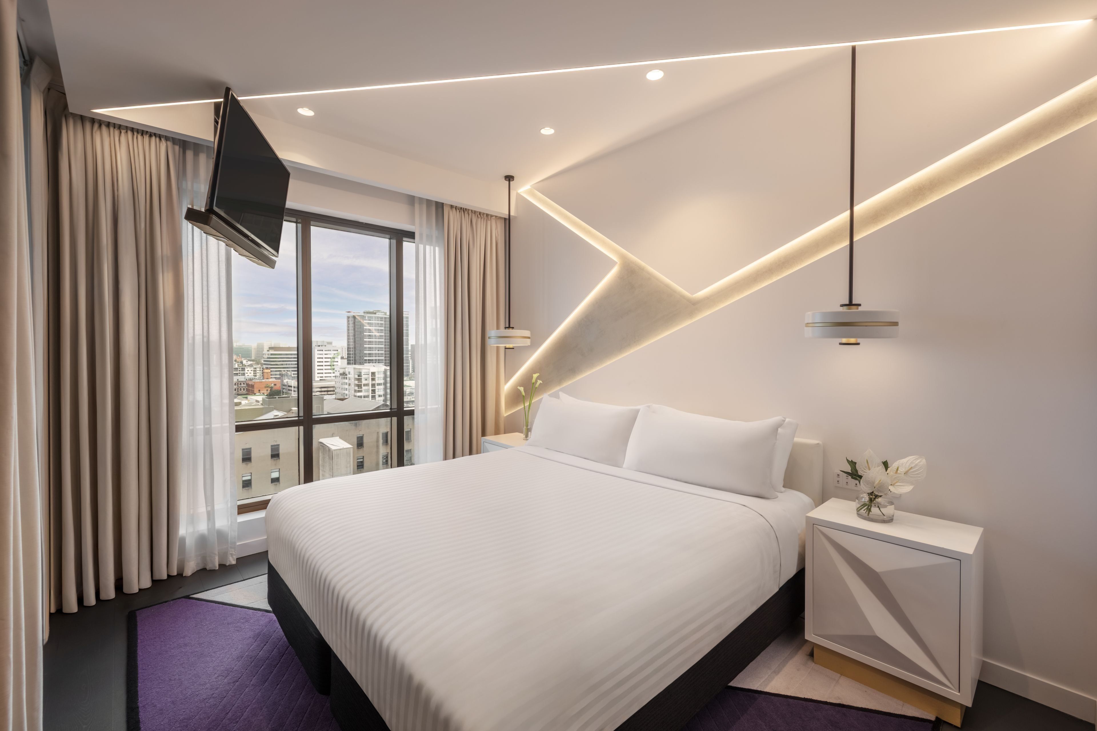 A modern hotel room with a large bed, a view of the city, and a unique geometric light design on the ceiling.