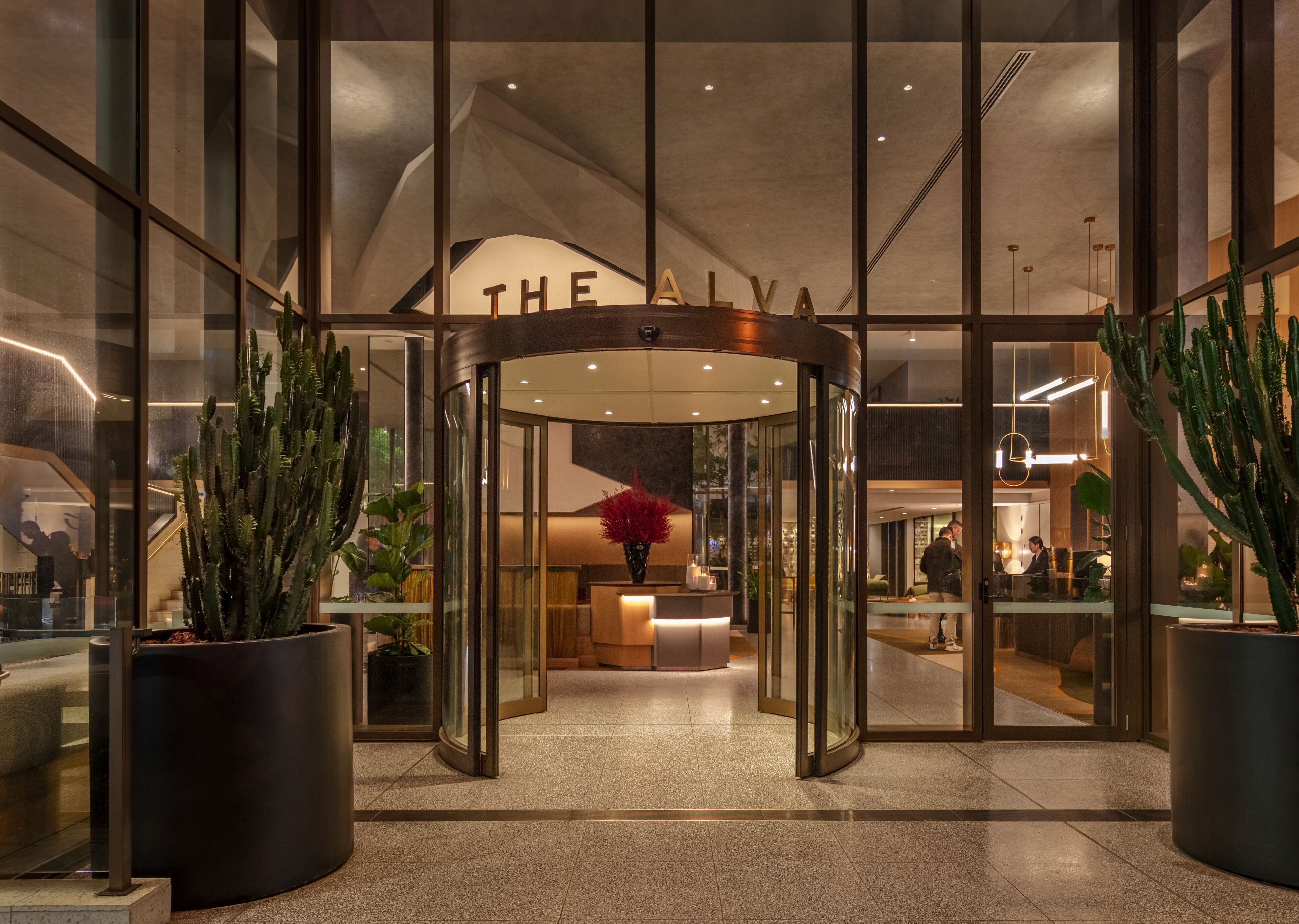 Entrada de un hotel con puerta giratoria, paredes de vidrio y cactus en macetas.
