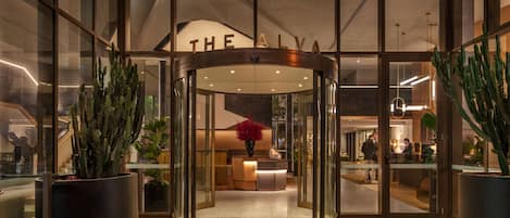 Entrada de un hotel con puerta giratoria, paredes de vidrio y cactus en macetas.