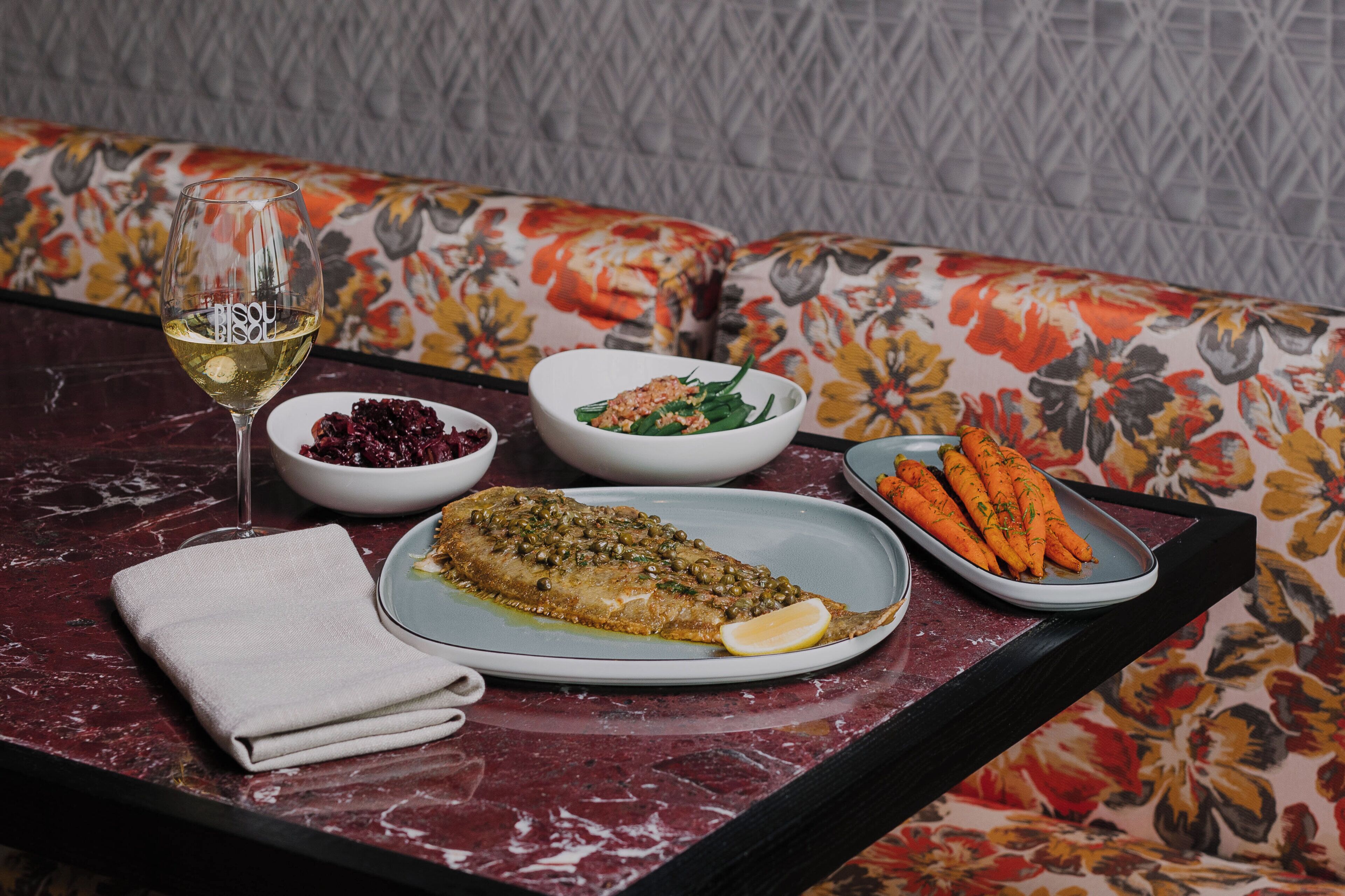 A dining table with a fish dish, salad, and carrots, a glass of white wine, and a folded napkin.