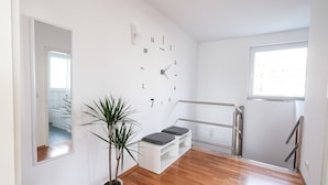 A modern, minimalistic room with a wooden floor, a potted plant, a bench with cushions, a wall clock, and a window.