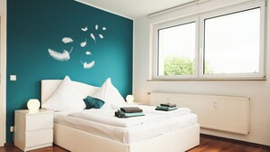 A modern bedroom with a teal accent wall, a white bed with pillows, a nightstand with a lamp, and a large window with a view of greenery.