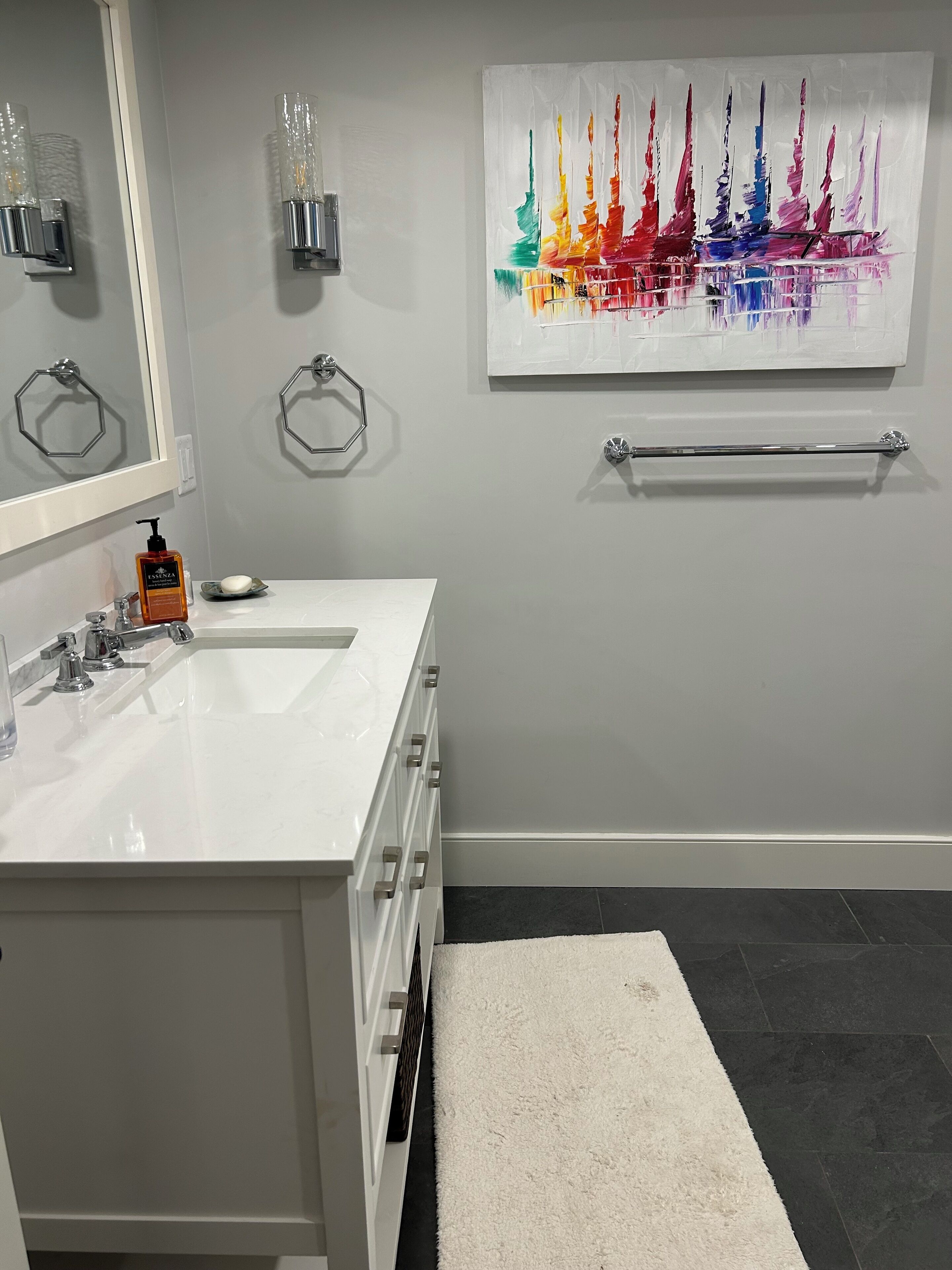 Un baño moderno con un tocador blanco, un espejo grande, un perchero y un cuadro abstracto de colores en la pared.