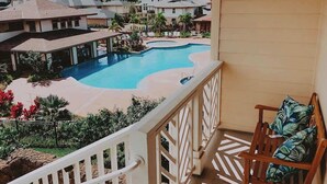 A pool area with a view of surrounding buildings, a wooden chair with cushions, and a balcony railing. - Pili Mai 7B - 4 Bedroom Suite in Poipu Sleeps 8 AC, Pool, Fitness Center & More! (Koloa)