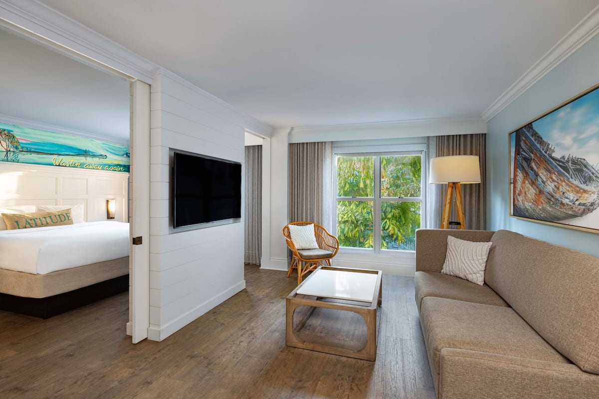 A hotel room with a bed, a sofa, a coffee table, a TV, a window with curtains, and a painting on the wall.