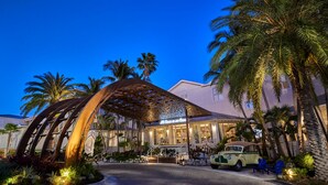 A hotel entrance with a palm tree-lined driveway, a vintage car, and a metal canopy structure.