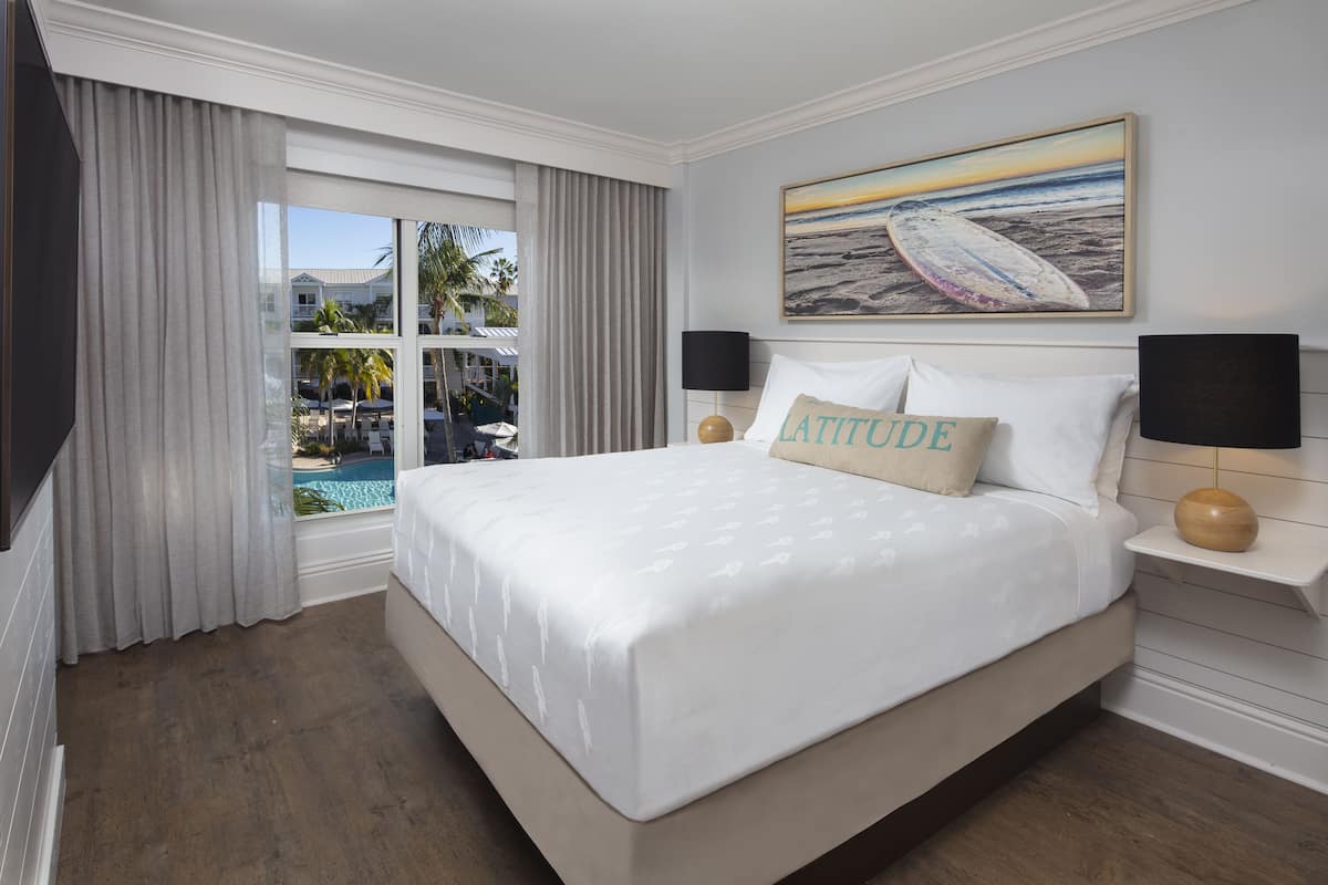 A hotel room with a large bed, a view of a pool and palm trees, and a painting of a beach scene.