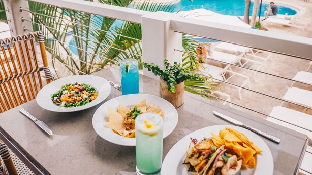 Una mesa de comedor con platos de comida, una bebida y vistas a una piscina y palmeras.