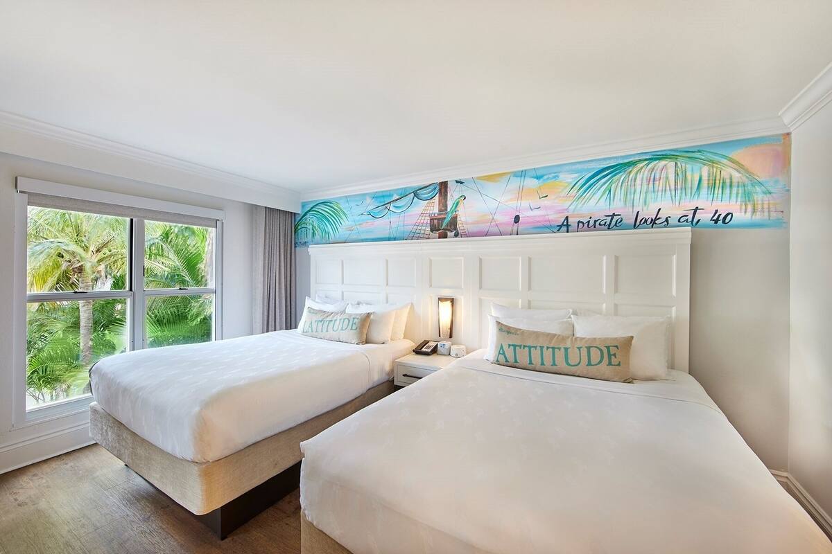 A hotel room with two beds, a large window with a view of palm trees, and a mural on the wall depicting a pirate ship.