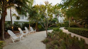 A sandy patio with three white Adirondack chairs, surrounded by lush tropical plants and a curved pathway.