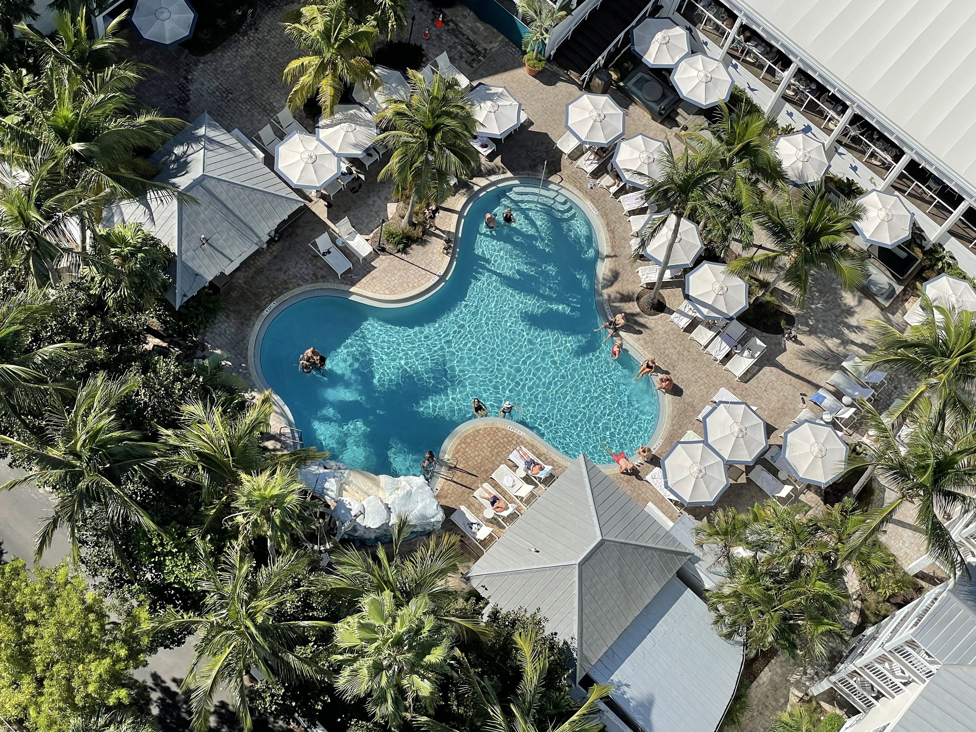 Vue aérienne d’un complexe hôtelier doté d’une piscine entourée de palmiers et de parasols blancs.