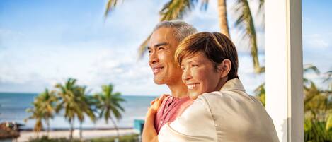 A couple embracing on a balcony with a view of the beach and palm trees.