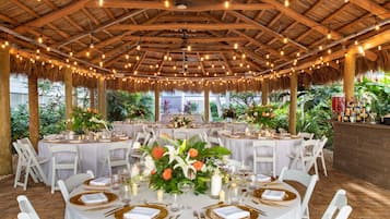 An outdoor dining area with round tables set for a meal, surrounded by white chairs and a thatched roof structure.