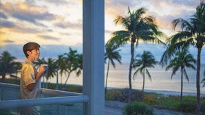 Una persona de pie junto a una ventana con vista a una playa, palmeras y el océano.